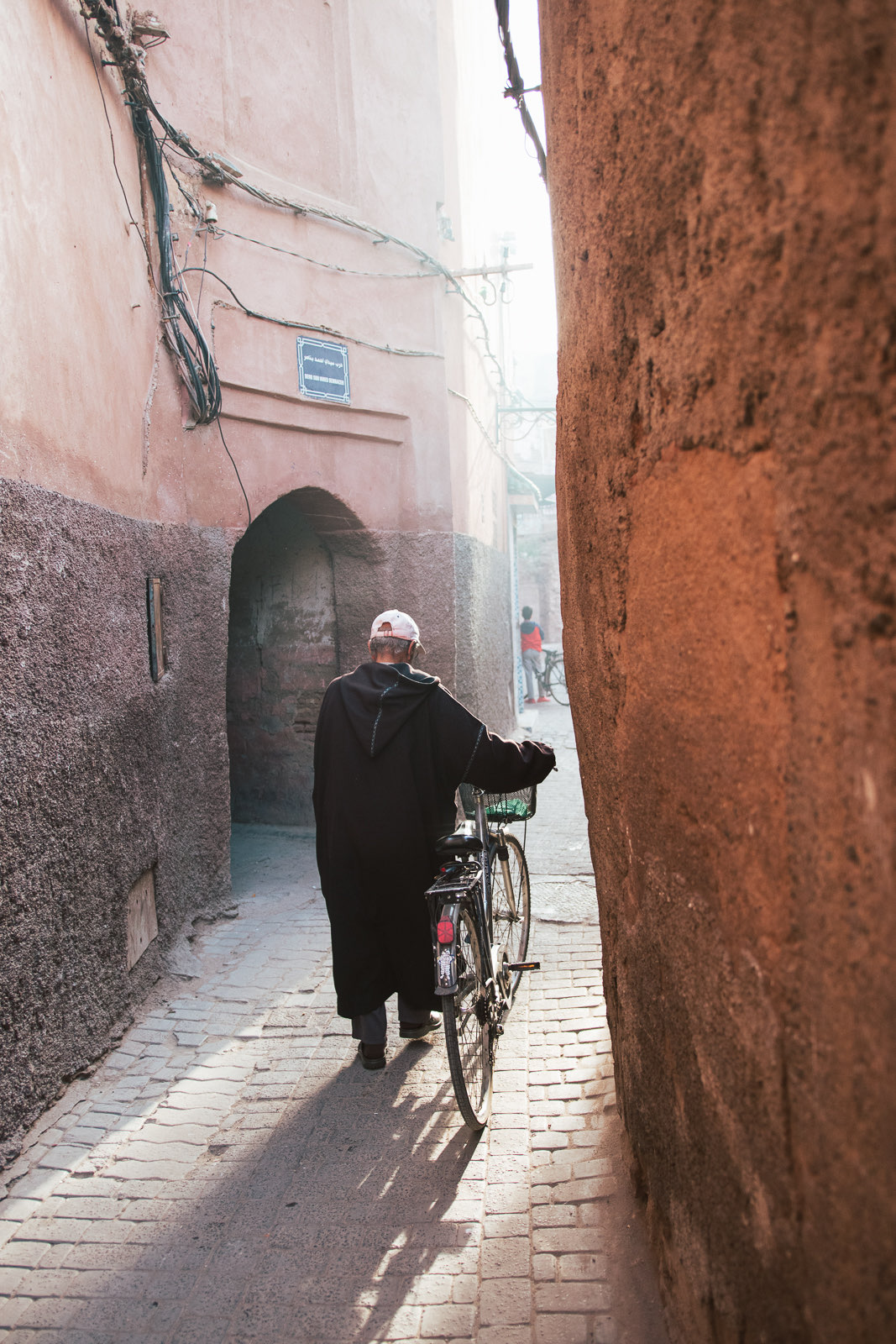 A man walking a bicycle through a narrow terracotta alley in the Marrakech medina